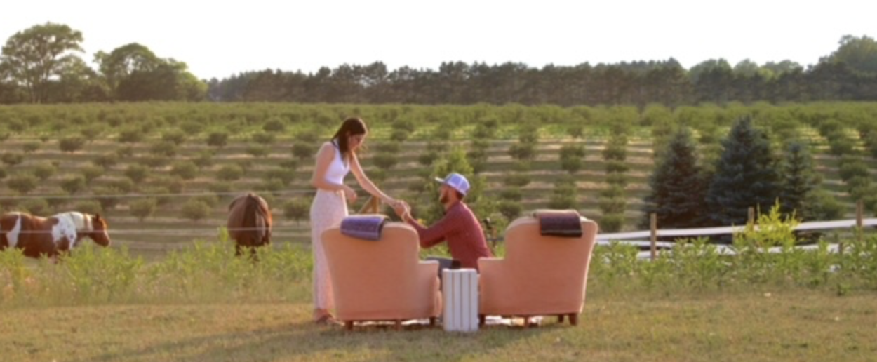 A young guy proposing to a woman in front of two orange chairs in a field